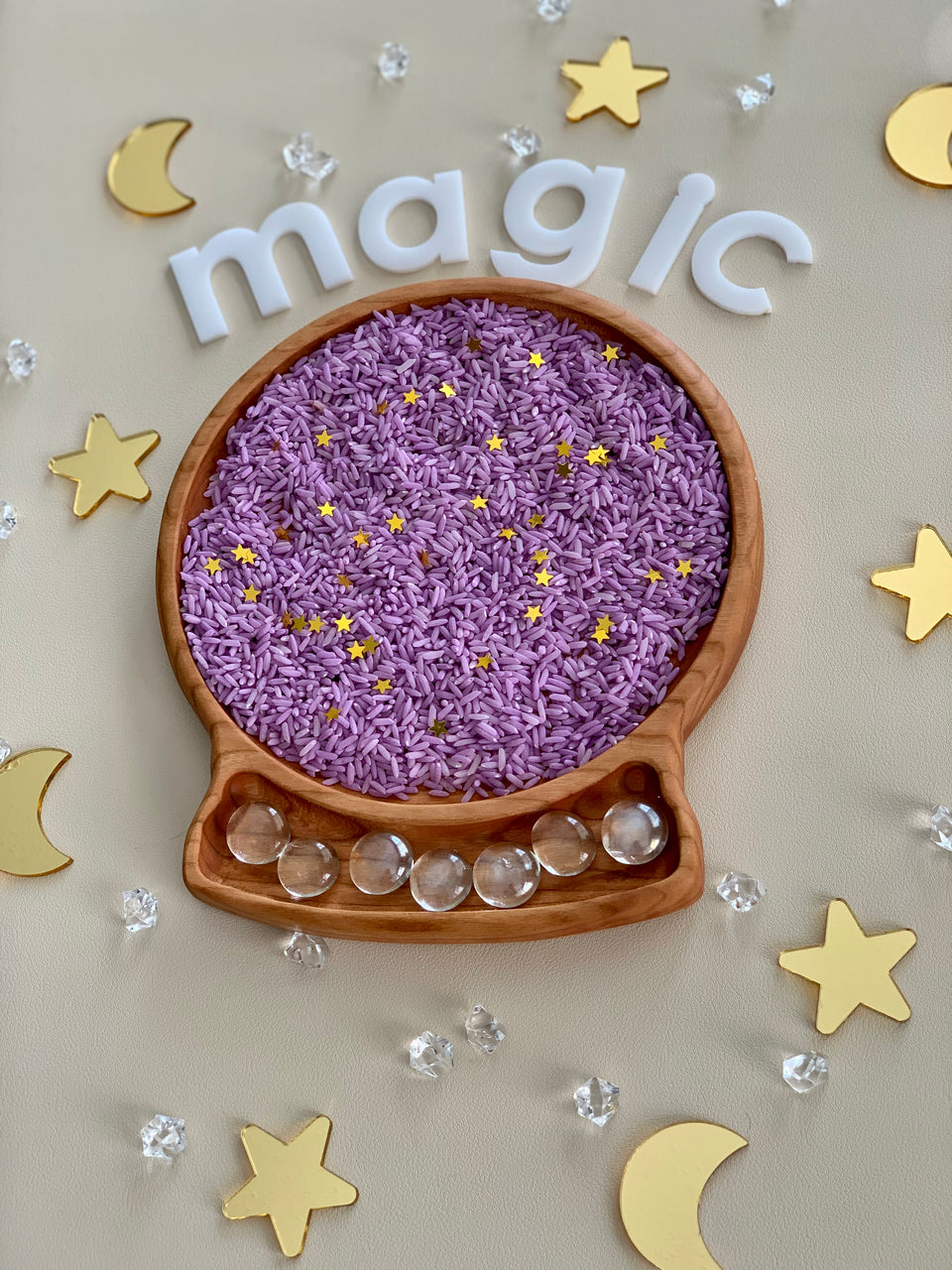 Wooden sensory tray shaped like a crystal ball filled with purple rice and gold star confetti on beige surface