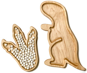 Natural wooden dinosaur footprint plate filled with small white beads next to empty dinosaur-shaped sensory tray