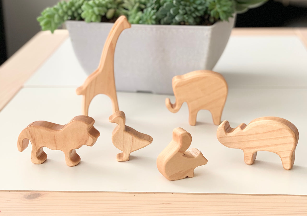 Natural wooden animal toy figurines including giraffe, elephant, lion, duck, squirrel, and rhino on white surface with plant background