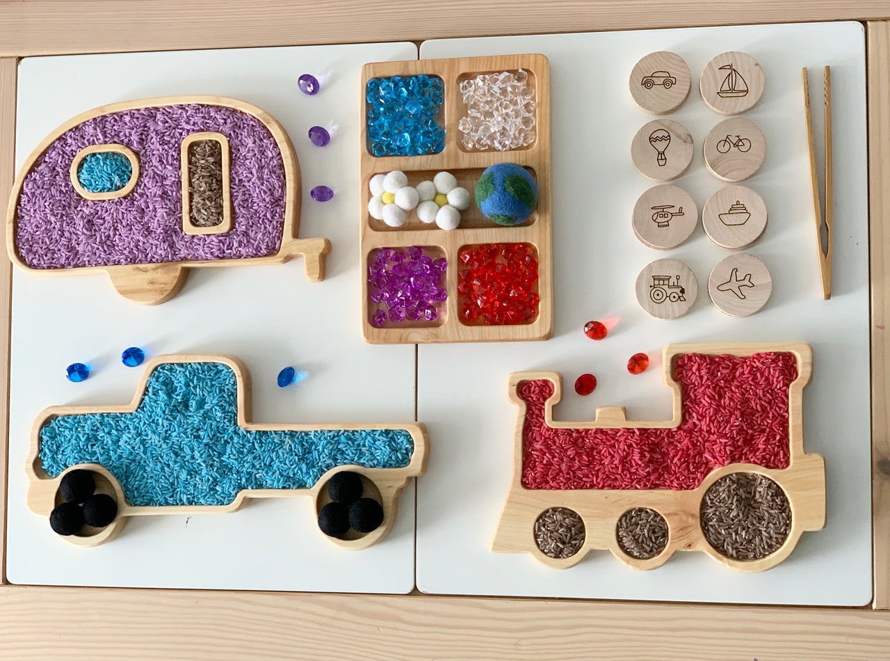 Wooden camper, truck, and train sensory trays filled with colored beads and textured materials on white surface with wooden tokens and tweezers