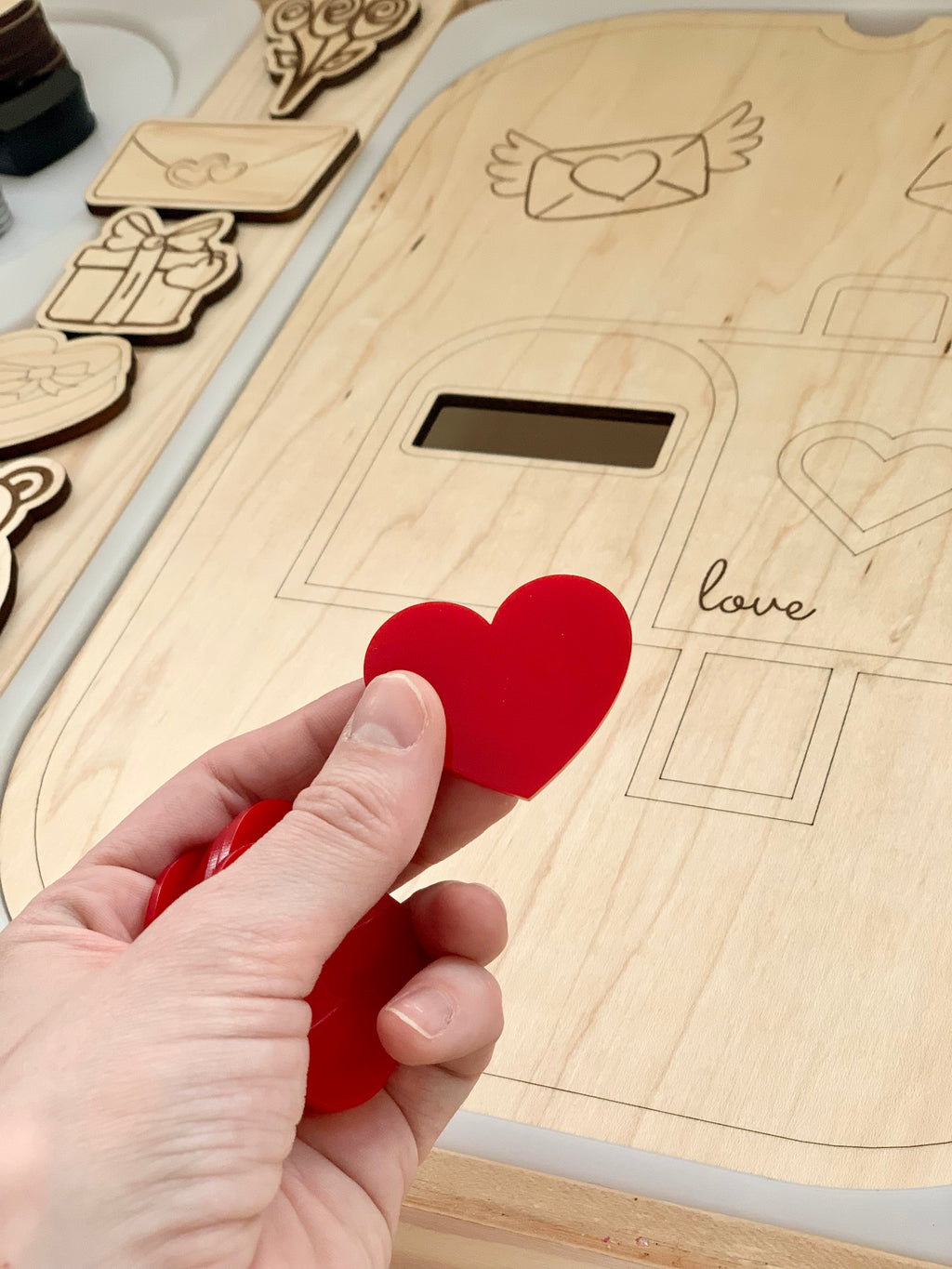 Hand holding red acrylic heart shape above wooden board with engraved heart and love letter shapes for sensory play
