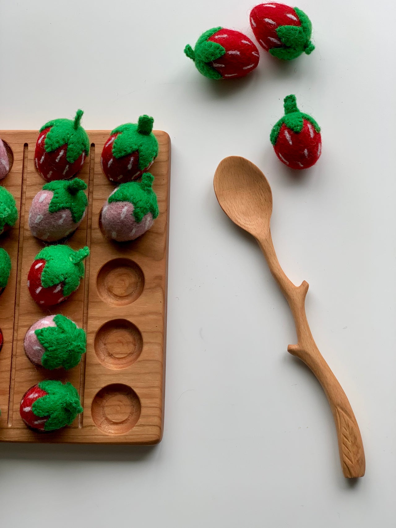 Natural wooden twig-shaped spoon with carved details next to handmade felt strawberries on white background