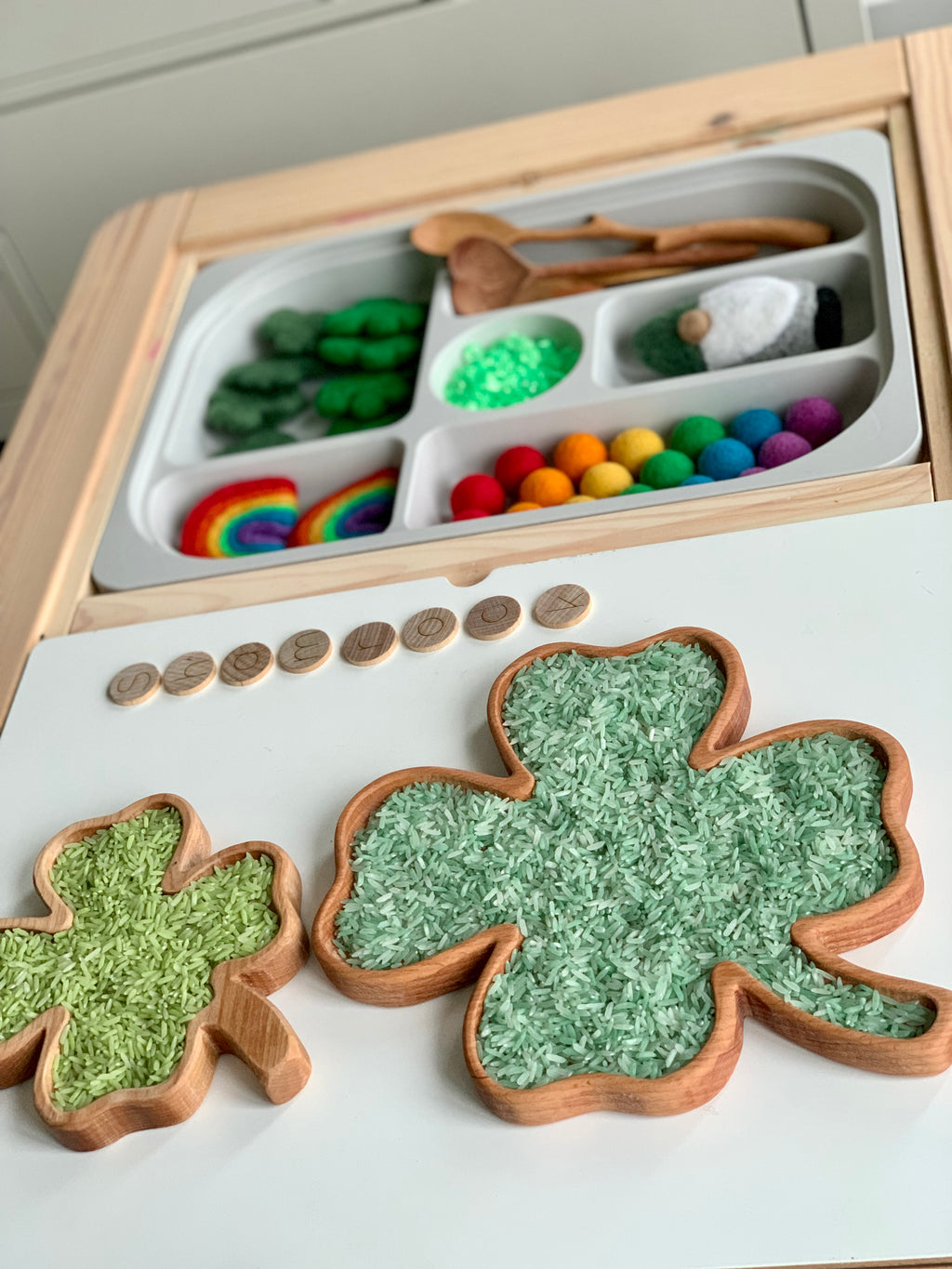 Small and large wooden shamrock sensory trays filled with green rice beside a white tray with wooden spoons and colorful felt balls