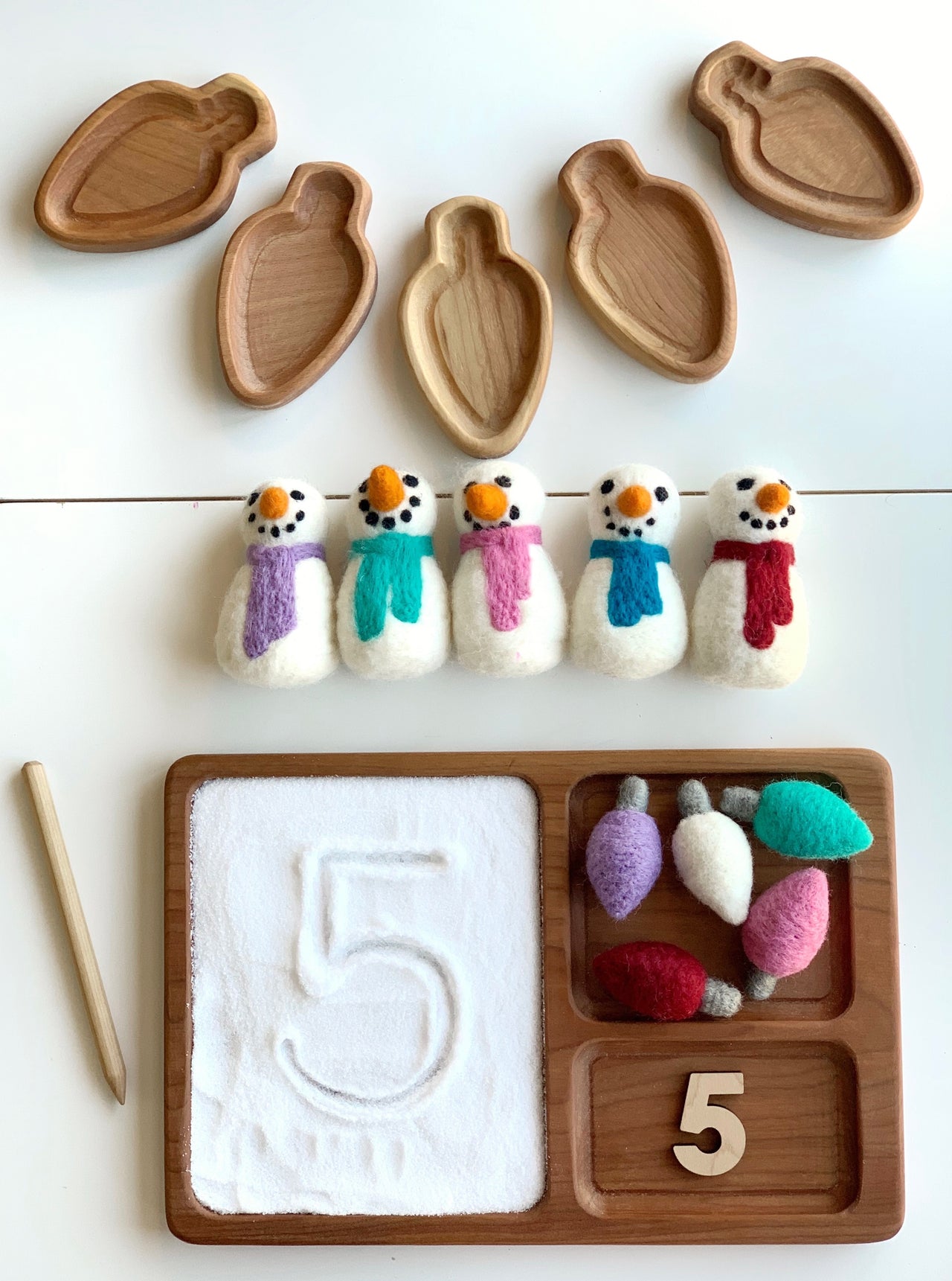 Natural wooden stylus with wooden tray containing white sand, felt snowmen, felt light bulbs, and number five cutout on white background