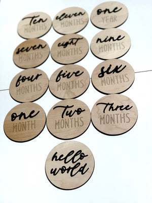 Natural wooden 3D milestone discs with black cursive and engraved letters displaying months and hello world text on white background