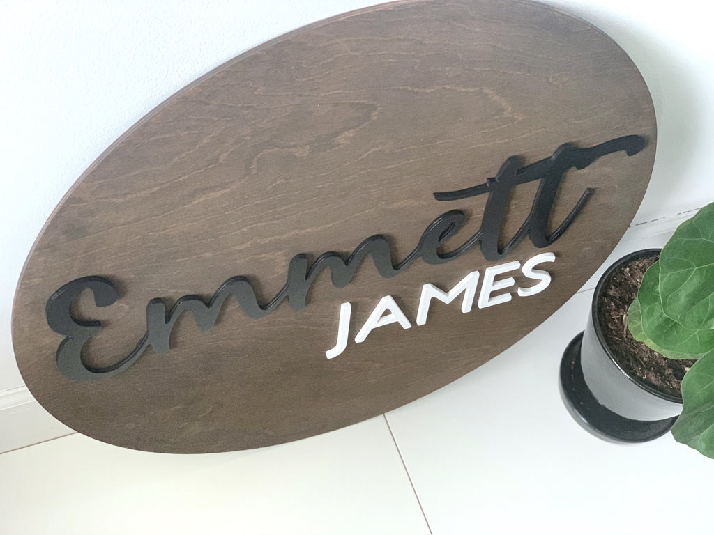 Oval custom wooden name sign with black cursive and white block letters on stained wood background next to potted plant