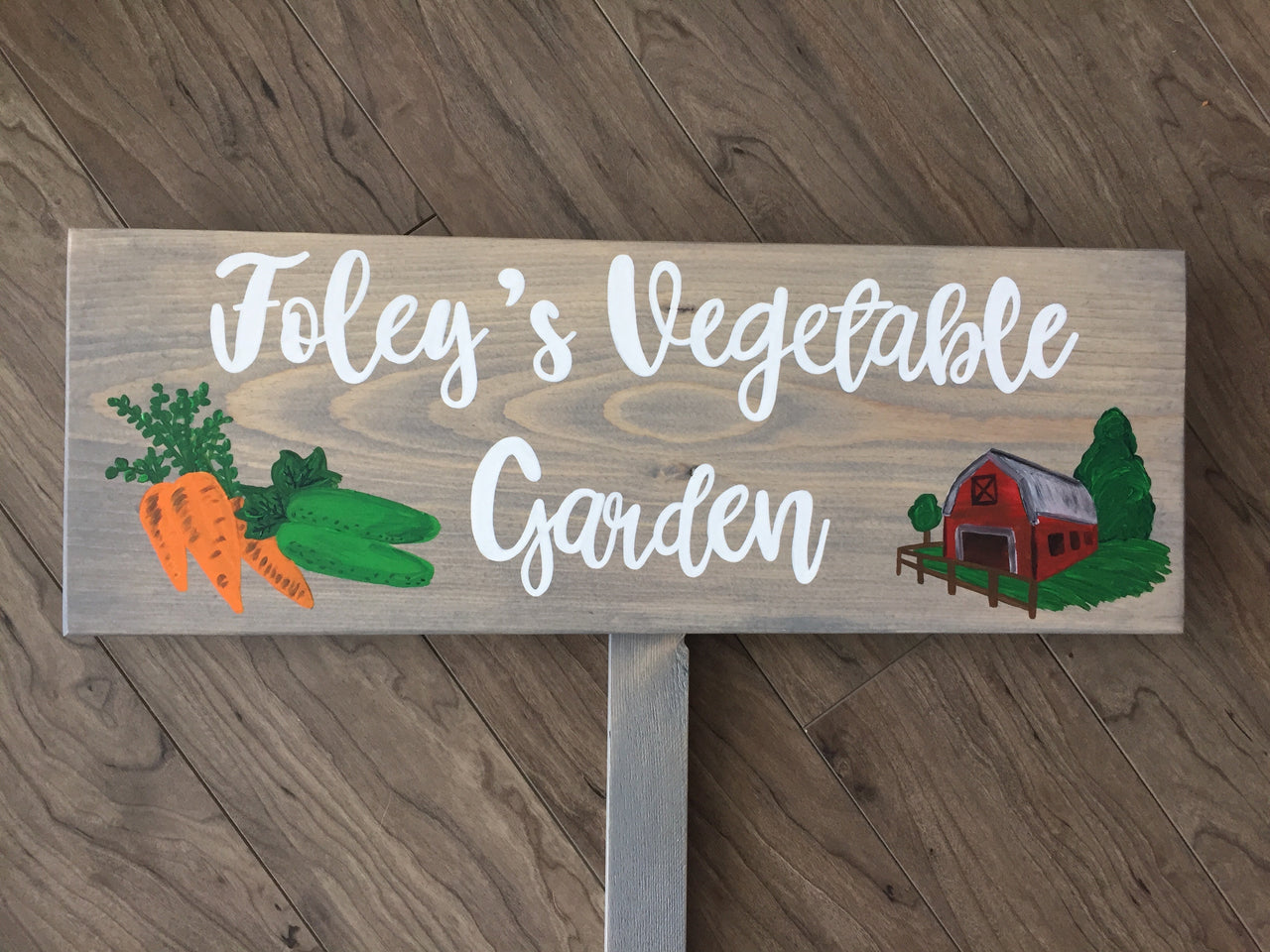 Large custom wooden vegetable garden sign with white cursive letters and carrot, cucumber, and red barn illustrations on wood background
