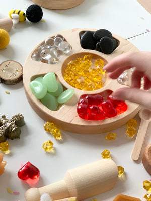 Round wooden tinker tray with compartments holding colorful translucent stones and a child's hand reaching in