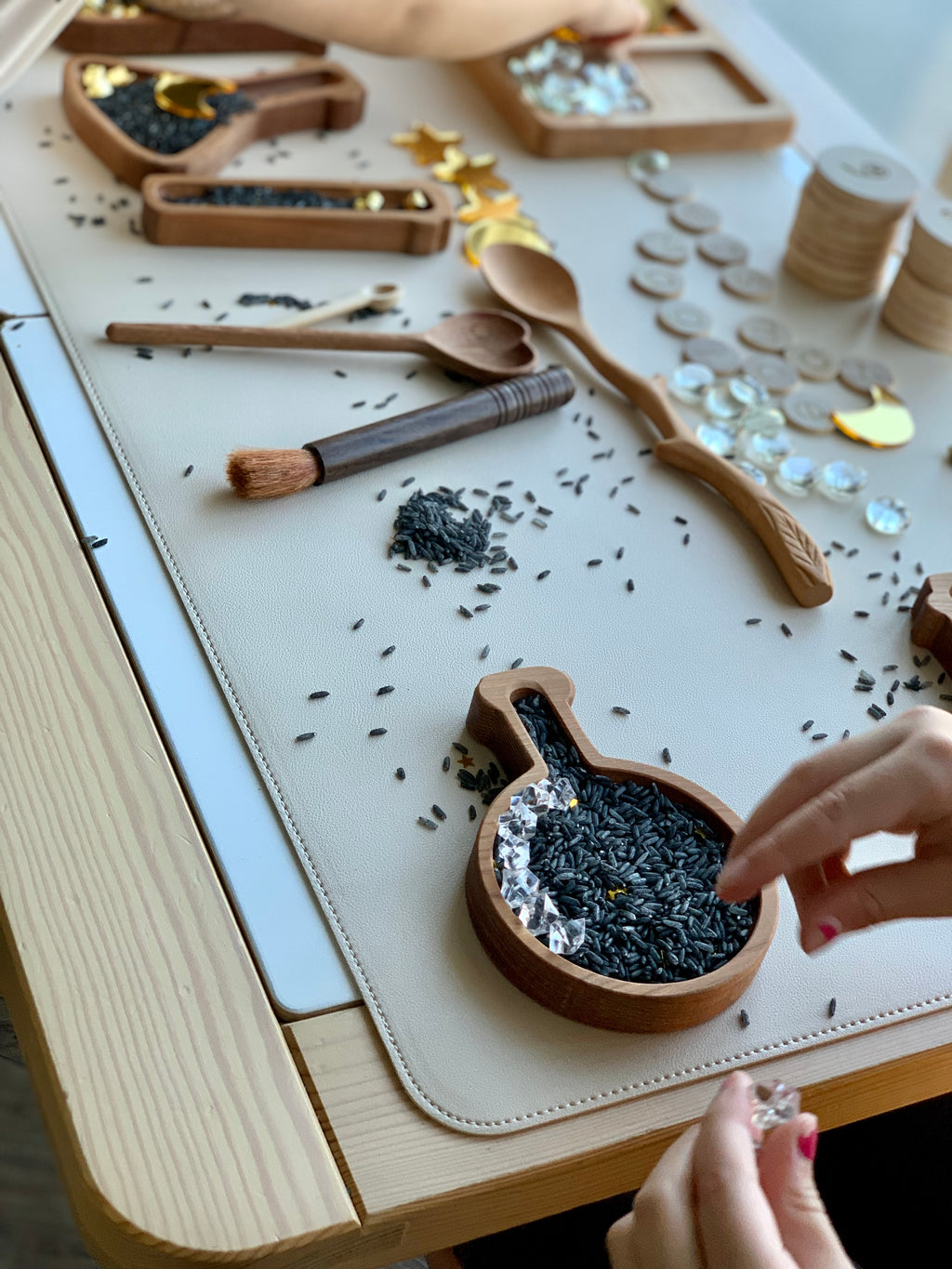 Twig style wooden spoon and wooden scoops with black rice and clear gems on beige mat with hands arranging gems