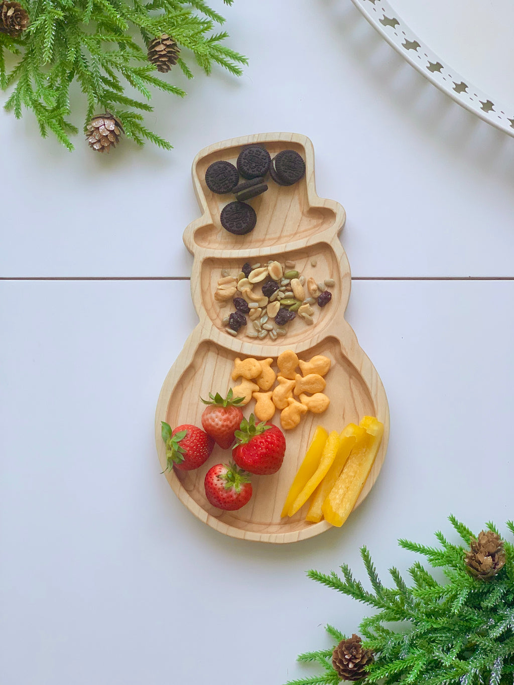 Natural wooden snowman-shaped plate with sections holding strawberries, goldfish crackers, yellow bell pepper, trail mix, and mini cookies