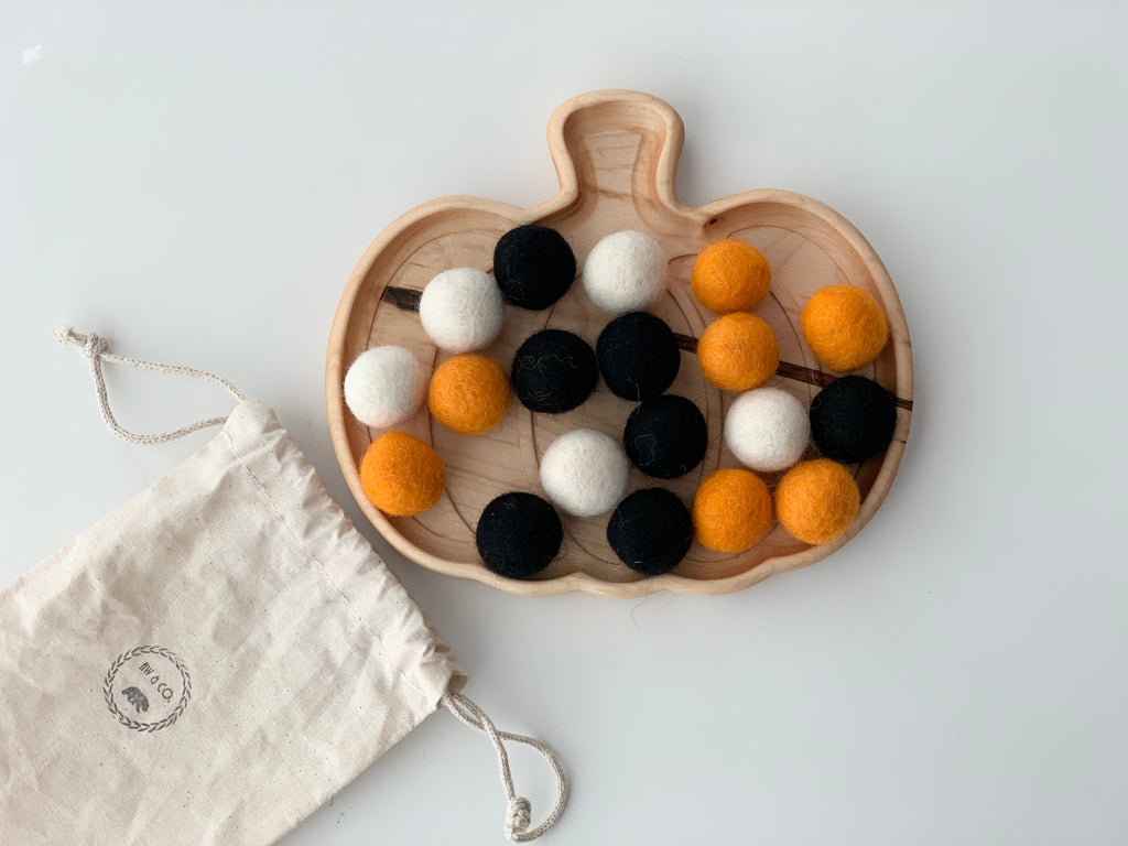 18 small felt balls in orange, black, and white on wooden pumpkin-shaped tray next to a drawstring pouch