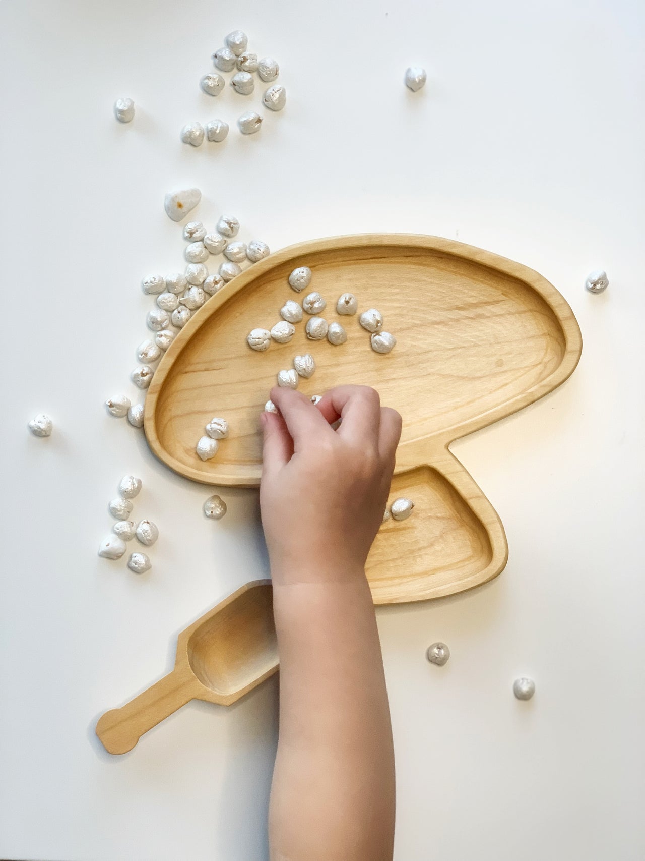 Natural wooden mushroom-shaped plate and small scoop with white pebbles scattered on white surface and child’s hand arranging pebbles
