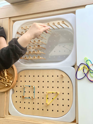 Child’s hand placing wooden peg into small hole geometric peg board with rubber band shapes on natural wood surface