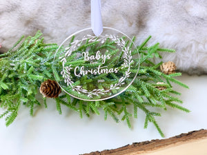Clear round acrylic baby's first Christmas ornament engraved with white lettering and leafy wreath on green pine background