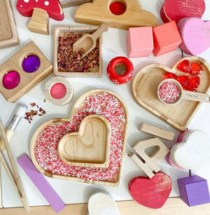 Natural wooden nesting heart plates filled with pink and white sensory materials surrounded by colorful wooden toys on white surface