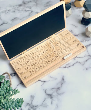 Wooden toy laptop with engraved keyboard keys and chalkboard screen on marble surface with decorative plants nearby