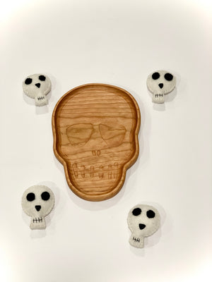 Natural wooden skull-shaped plate with engraved features surrounded by four small white felt skull toys on white background