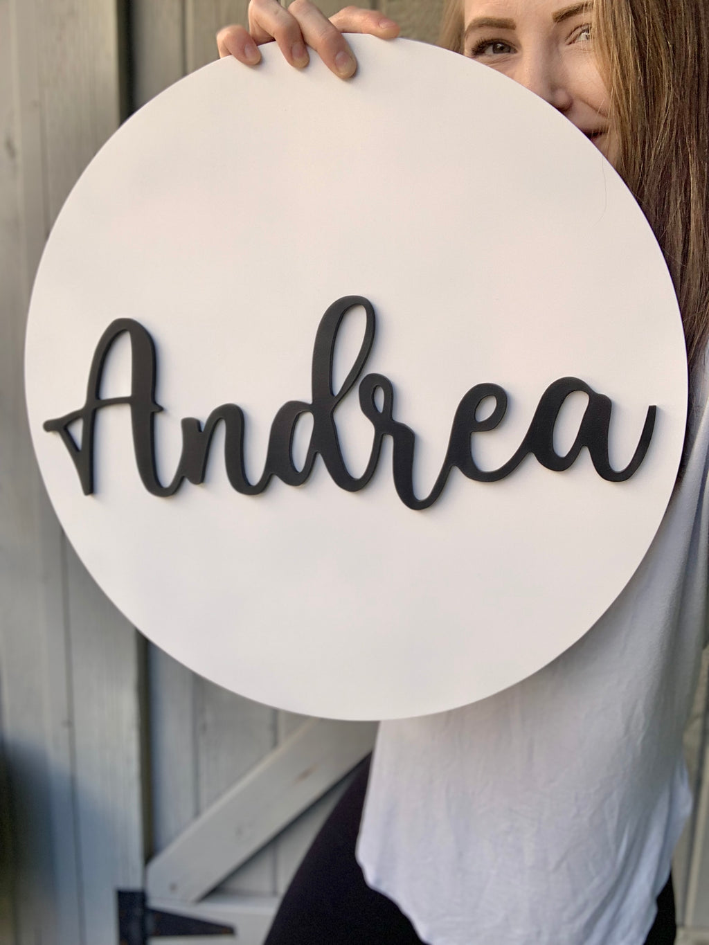 Round white 3D name sign with black cursive letters spelling Andrea held by person in light gray shirt against wooden background