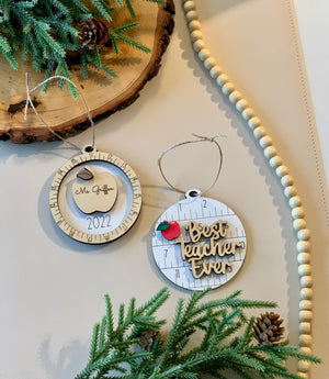Two round wooden Christmas ornaments with teacher-themed designs hanging on a beige surface with greenery and pinecones nearby