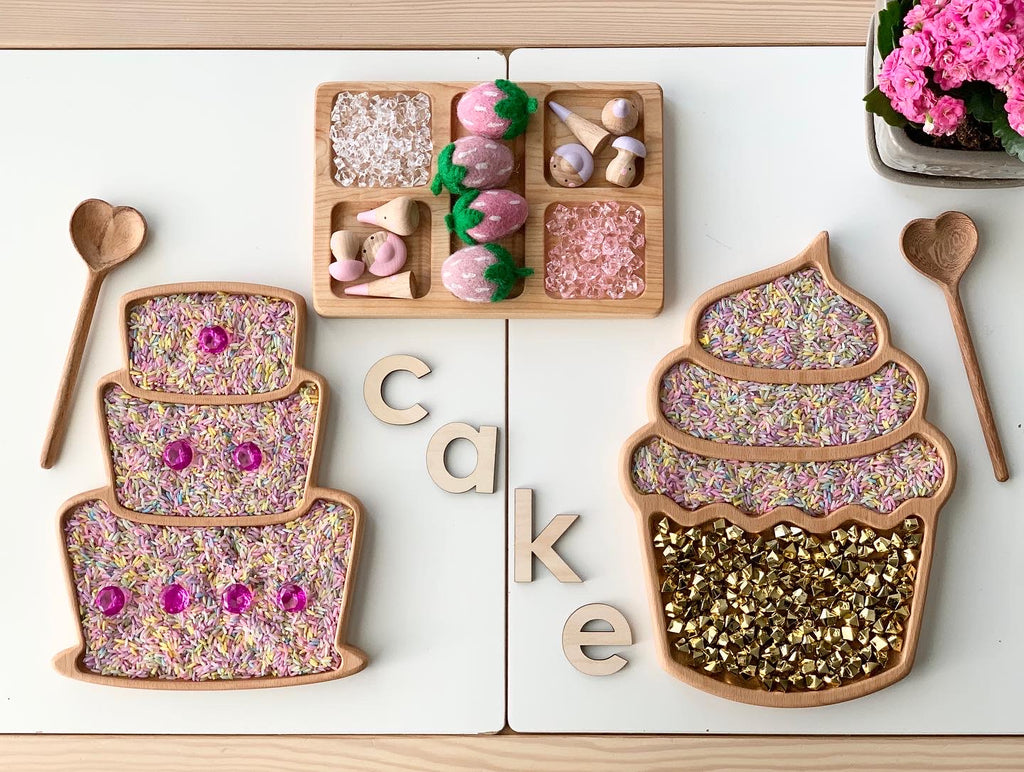 Wooden cupcake and cupcake-shaped sensory trays filled with colorful dried flowers and gold beads on white surface