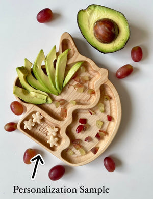 Natural wooden unicorn-shaped plate with engraved name sample, sliced avocado, grapes, and small cereal pieces on white surface