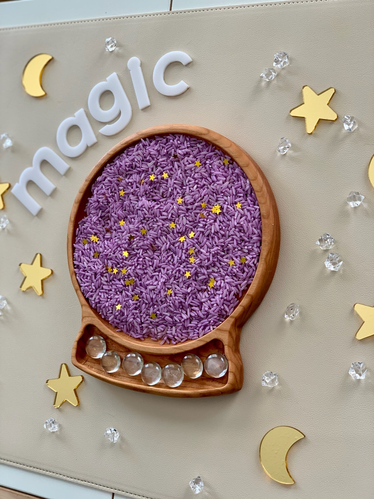 Wooden tray shaped like a crystal ball filled with purple dyed rice and scattered gold star confetti on beige surface