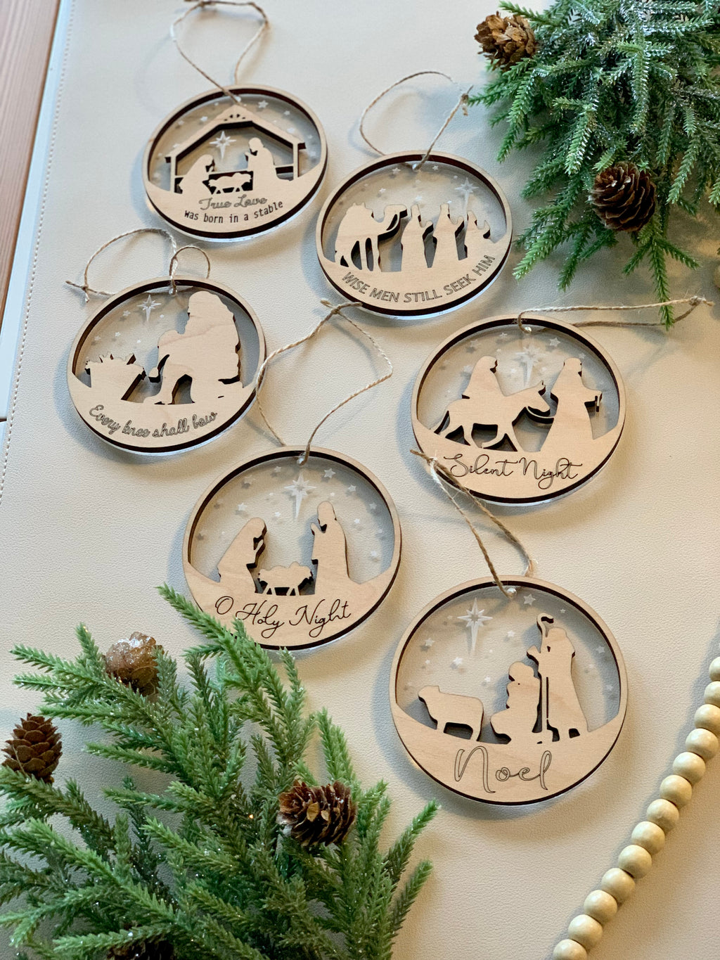 Two-layer wood and acrylic nativity Christmas ornaments with biblical scenes and festive inscriptions on white surface with greenery