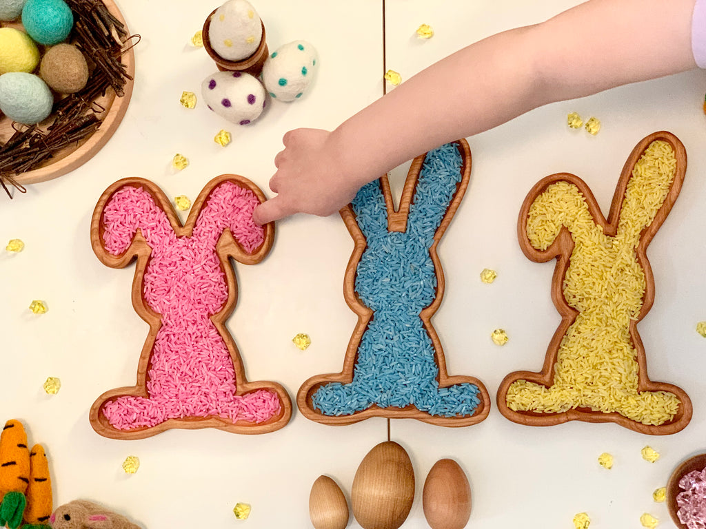 Three wooden bunny-shaped sensory trays filled with colorful rice and a child's hand reaching to touch one on white table