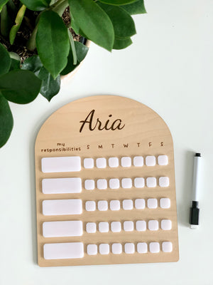 Personalized wooden chore chart with white acrylic dry erase pieces and black marker on white surface next to green plant