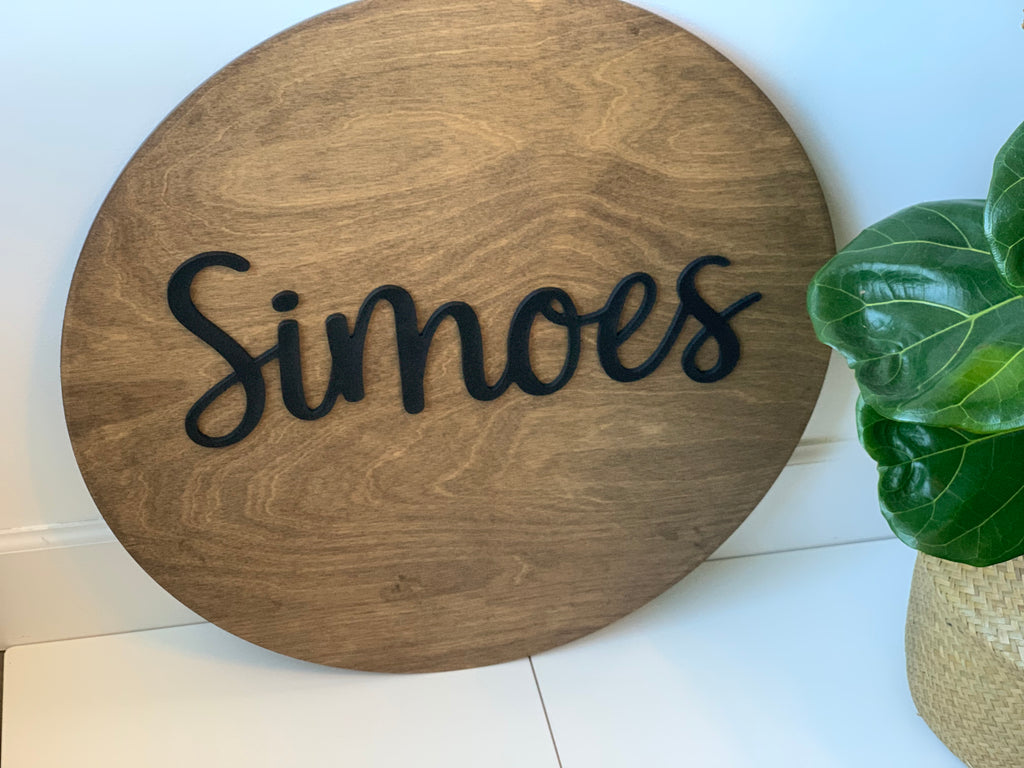 Round wooden name sign with black cursive letters on natural wood background beside green potted plant