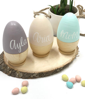 Three personalized hollow fillable wooden Easter eggs in gray, cream, and blue on rustic wood slice with candy eggs