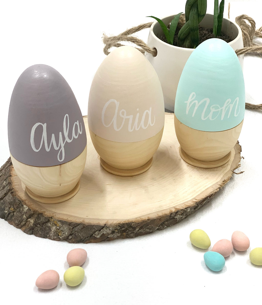 Three personalized hollow fillable wooden Easter eggs in gray, cream, and blue on rustic wood slice with candy eggs