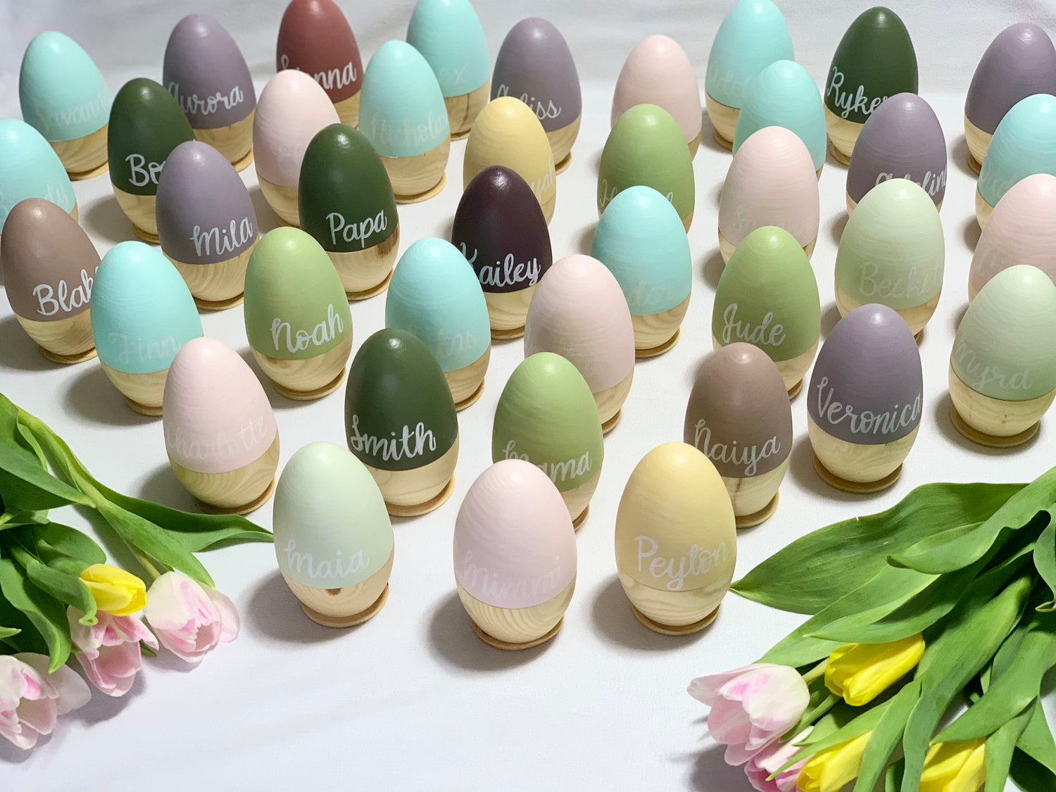 Rows of pastel-colored hollow fillable wooden Easter eggs with white personalized names on natural wood stands and tulips nearby
