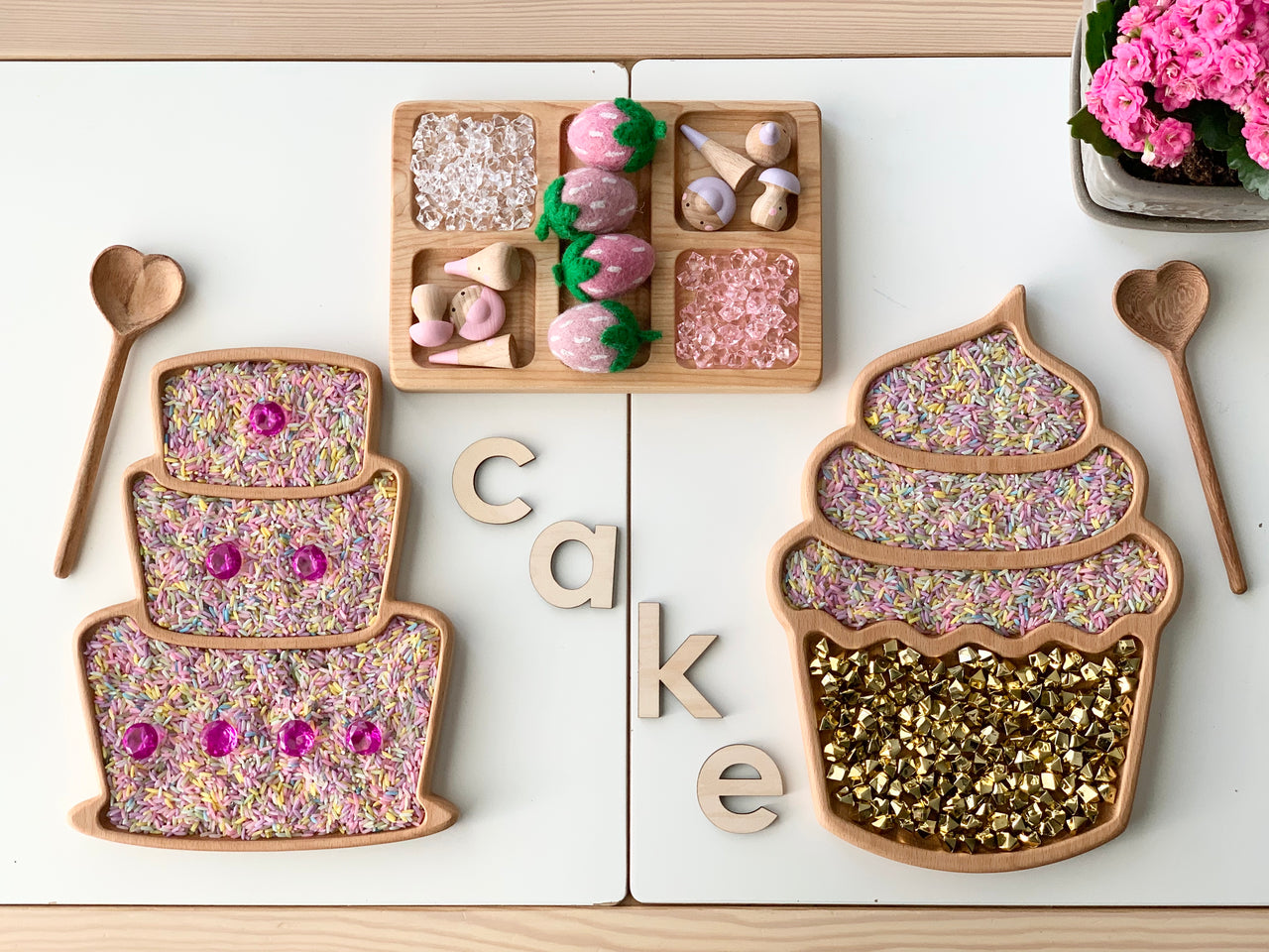Wooden cupcake and cupcake-shaped sensory trays filled with colorful rice, flower petals, and golden beads with wooden spoons and letters spelling cake