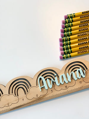 Natural wooden personalized ruler with engraved rainbow and cloud designs spelling Ariana on white background with pencils
