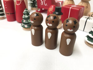 Three handmade wooden reindeer peg dolls with painted antlers and white chest spots on white surface with holiday decorations