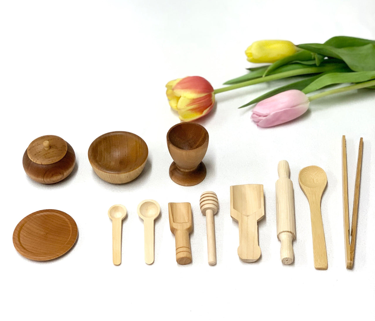 Small wooden sensory bin tools including bowls, spoons, rolling pin, honey dipper, and chopsticks on white background with tulips