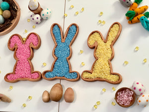 Three wooden bunny-shaped sensory trays filled with pink, blue, and yellow rice surrounded by Easter decorations on white table