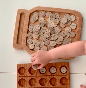 Wooden 1-inch alphabet and number discs with simple laser engraved letters and numbers on natural wood tray with child’s hand