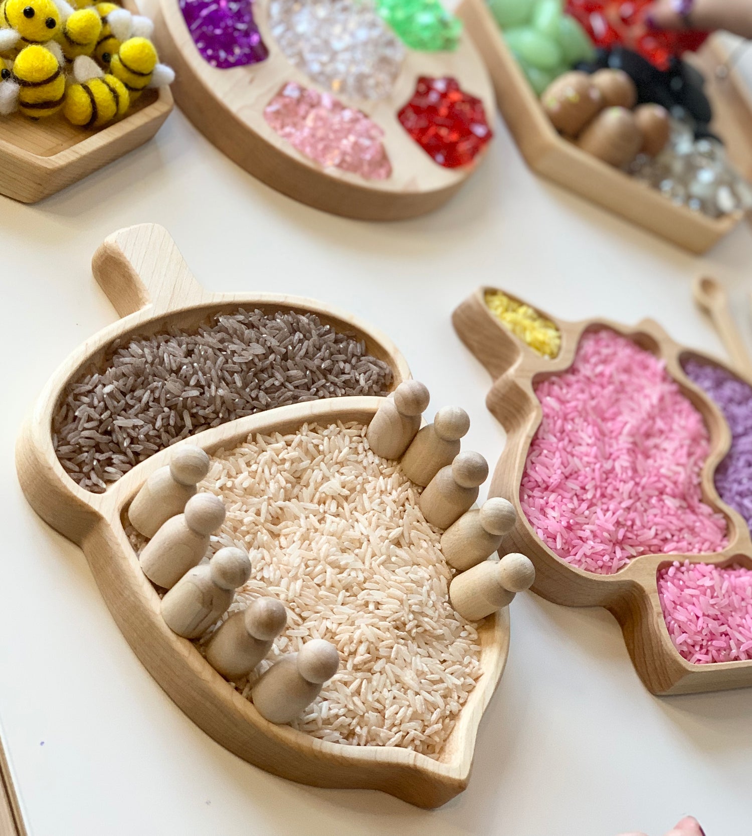 Wooden acorn-shaped sensory tray filled with dyed and natural rice and small wooden peg dolls on white background