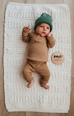 Infant lying on textured white blanket wearing brown outfit and green hat with one month wooden milestone disc nearby