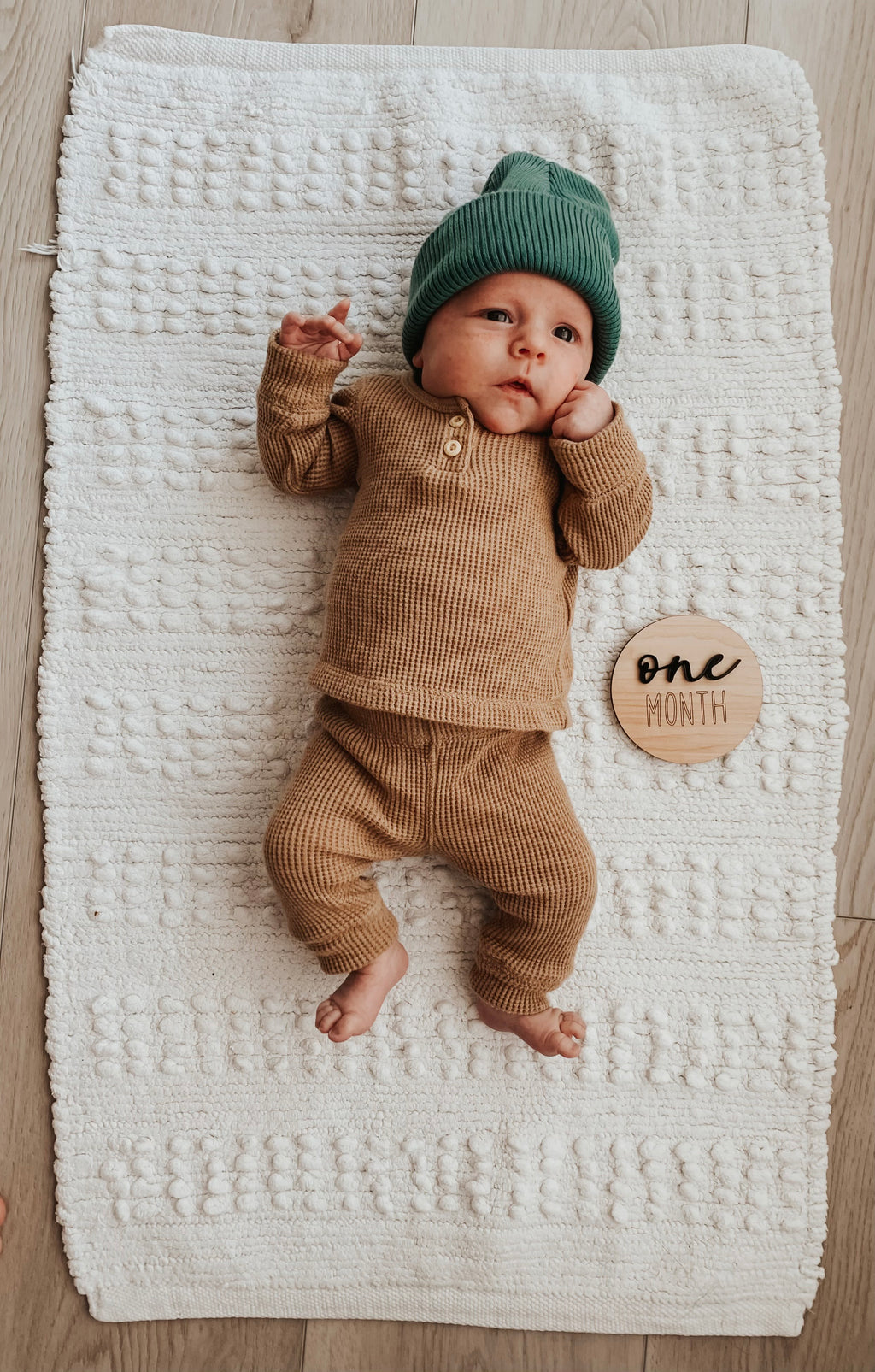 Infant lying on textured white blanket wearing brown outfit and green hat with one month wooden milestone disc nearby