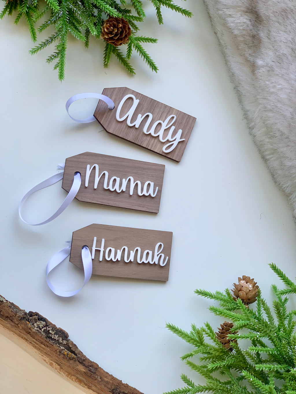 Set of three custom wooden Christmas stocking tags with white cursive names and white ribbon on white surface with green pine accents