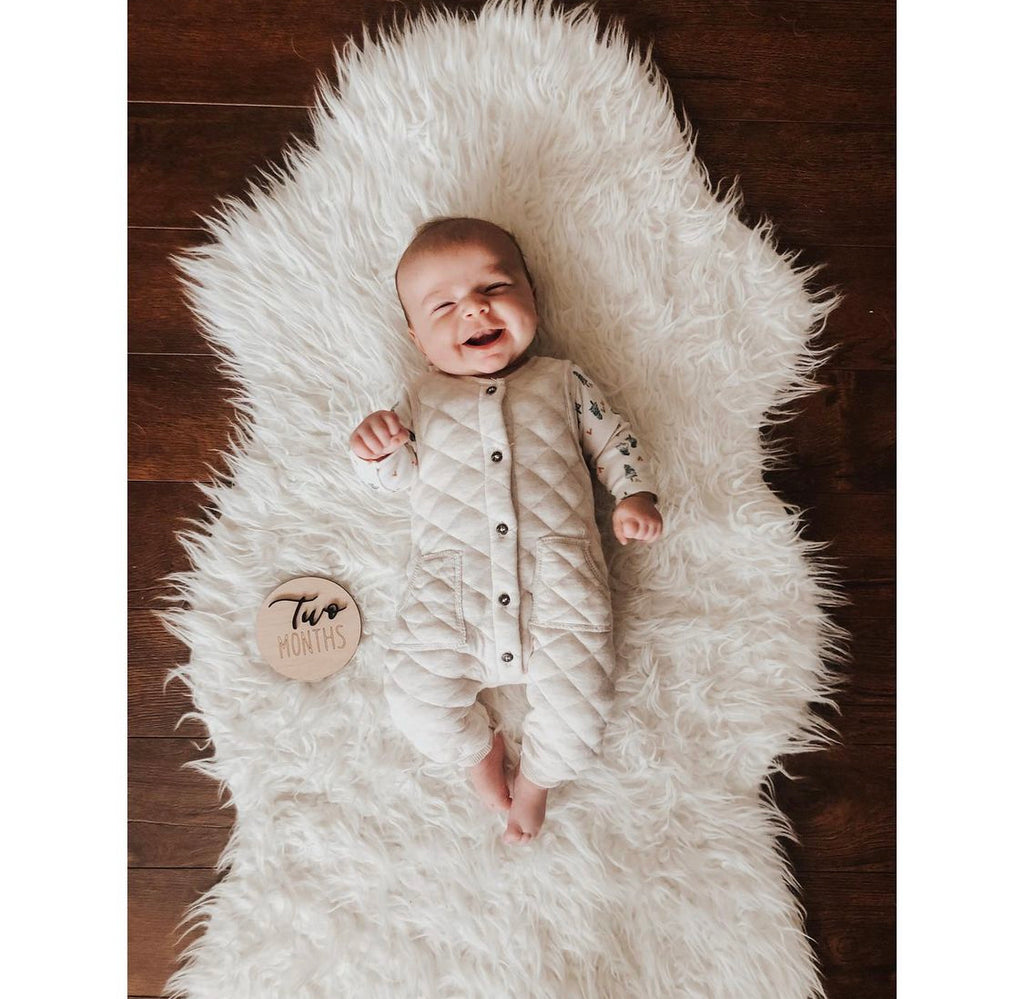 Smiling baby lying on white fluffy rug next to wooden disc engraved with two months milestone text