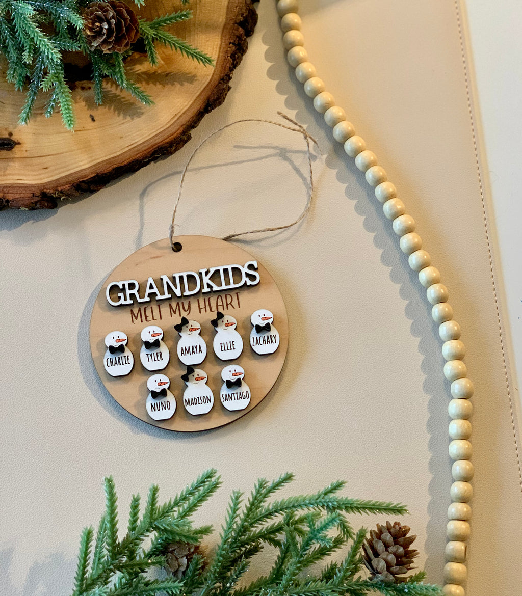 Round wooden Christmas ornament with snowman figures and names hanging on twine with greenery and wooden beads nearby