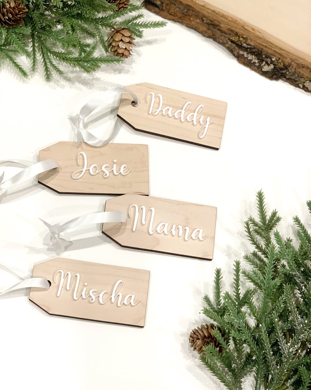 Custom wooden gift tags with white cursive names and white ribbons surrounded by pine branches and pine cones on white surface