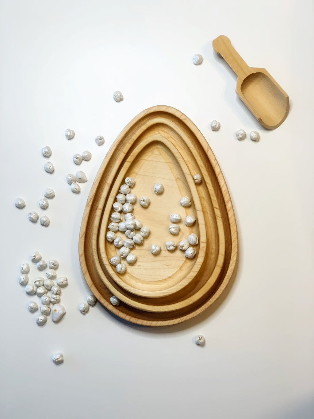 Set of natural wooden nesting egg plates with white sensory pebbles and small wooden scoop on white surface