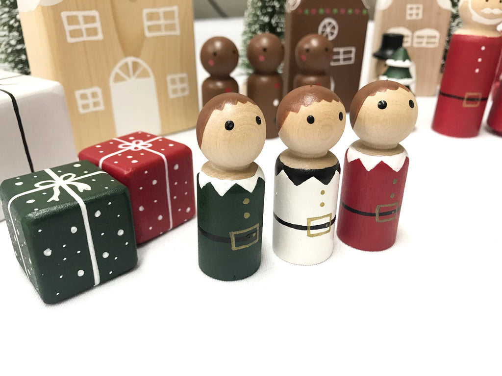 Three wooden peg dolls dressed as Christmas elves with painted green, white, and red outfits beside two wrapped gift blocks