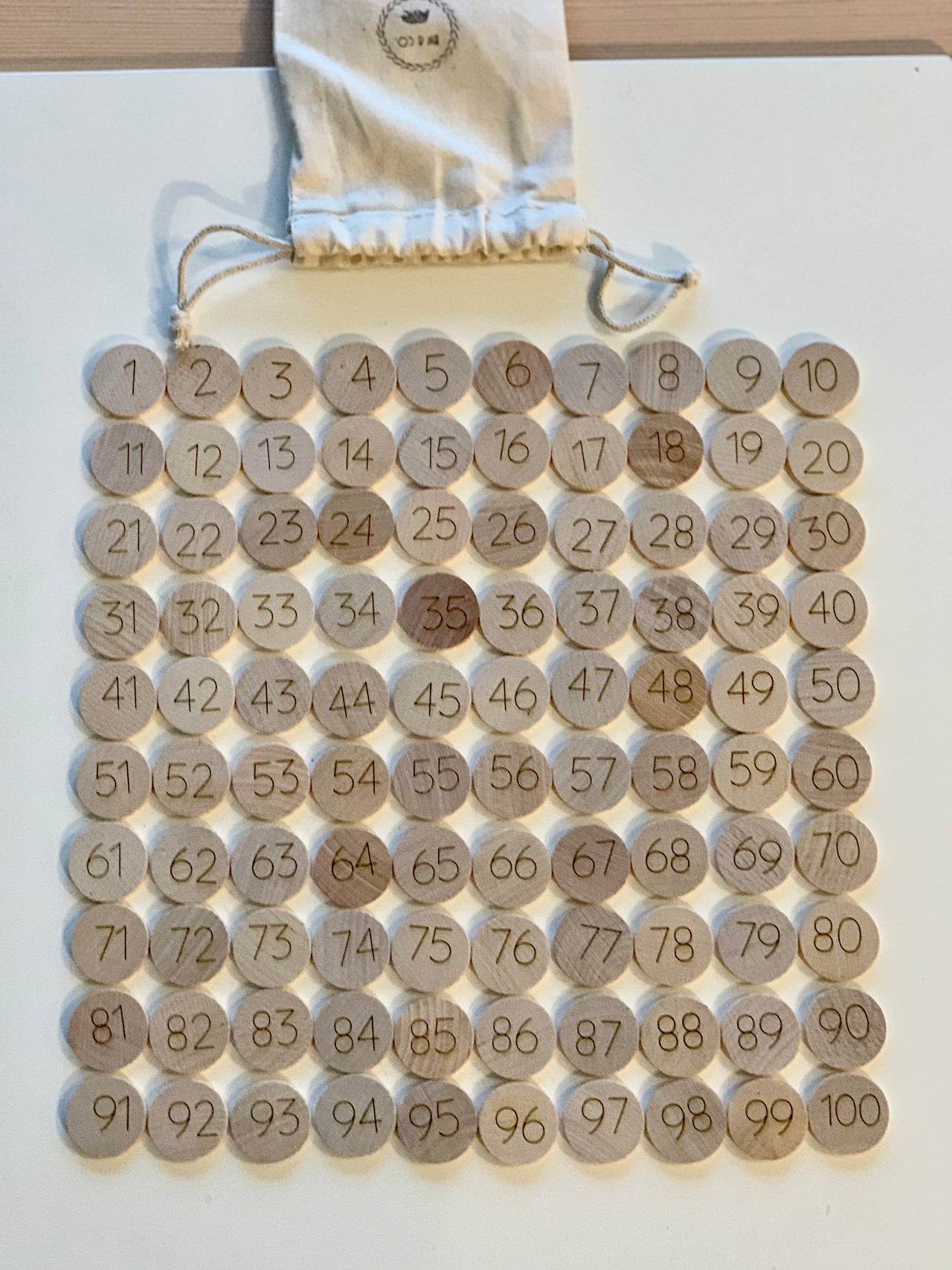 Set of 1-100 laser engraved wooden number discs neatly arranged next to drawstring cloth bag on white surface
