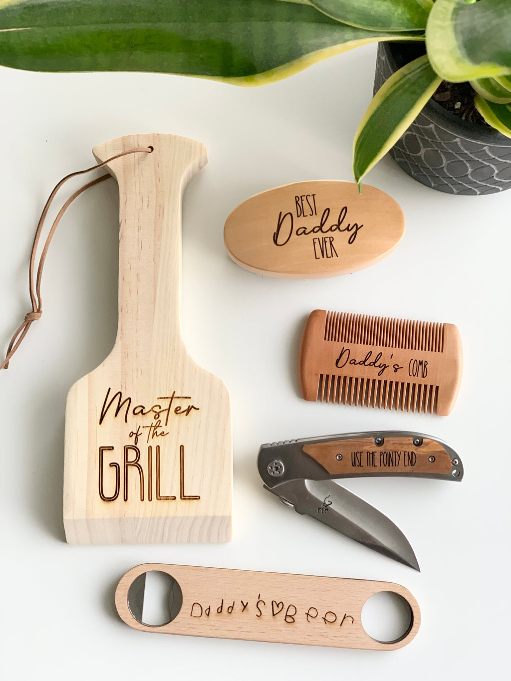 Set of personalized wooden gifts including paddle spatula, comb, oval box, bottle opener, and folding pocket knife on white surface